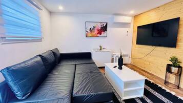 Un salon moderne avec un canapé en cuir noir, une table basse blanche, un téléviseur fixé au mur et un petit meuble blanc.