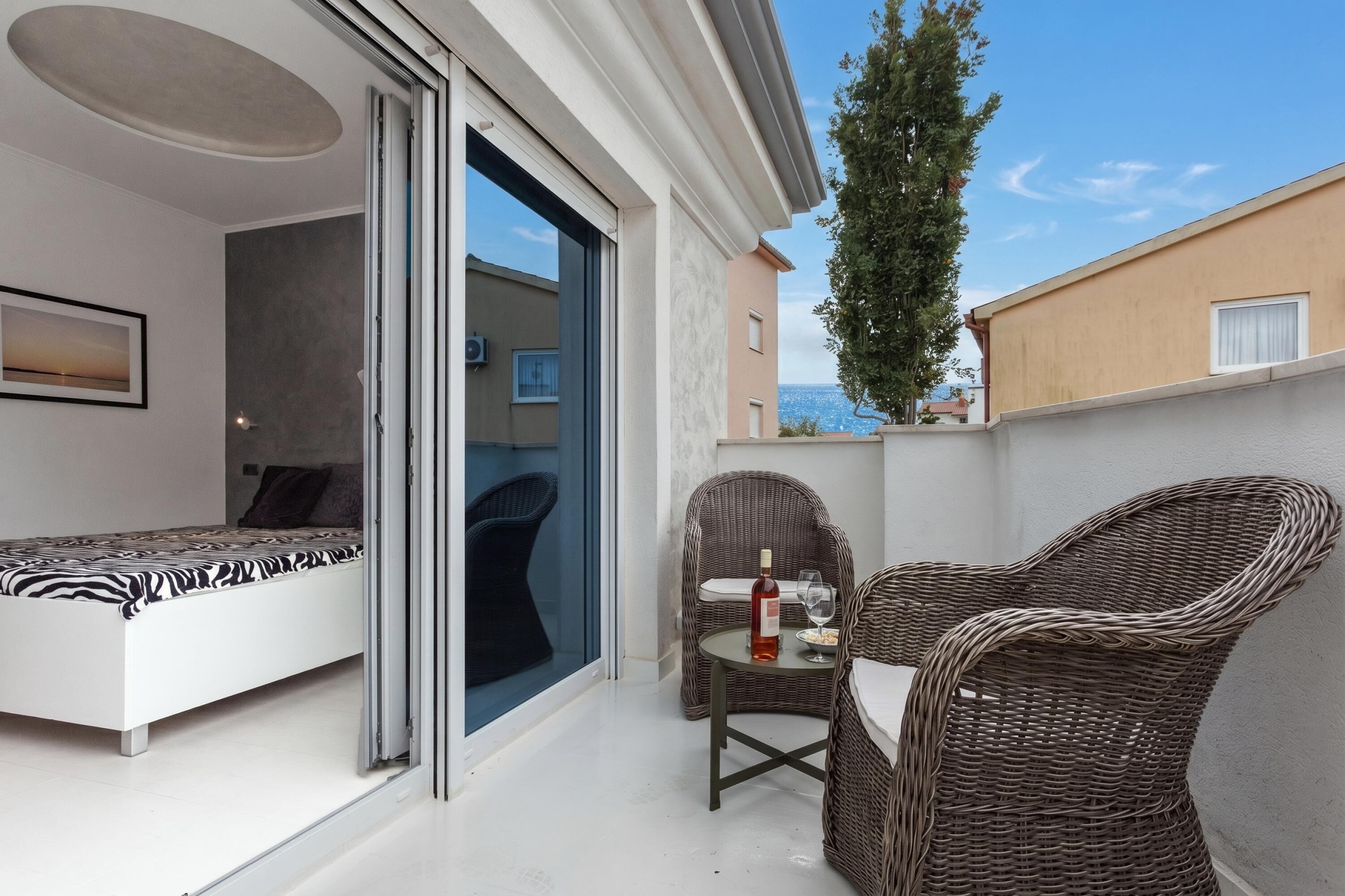 Een balkon met een bed, rieten stoelen, een tafel met een fles en een glas, en uitzicht op zee.