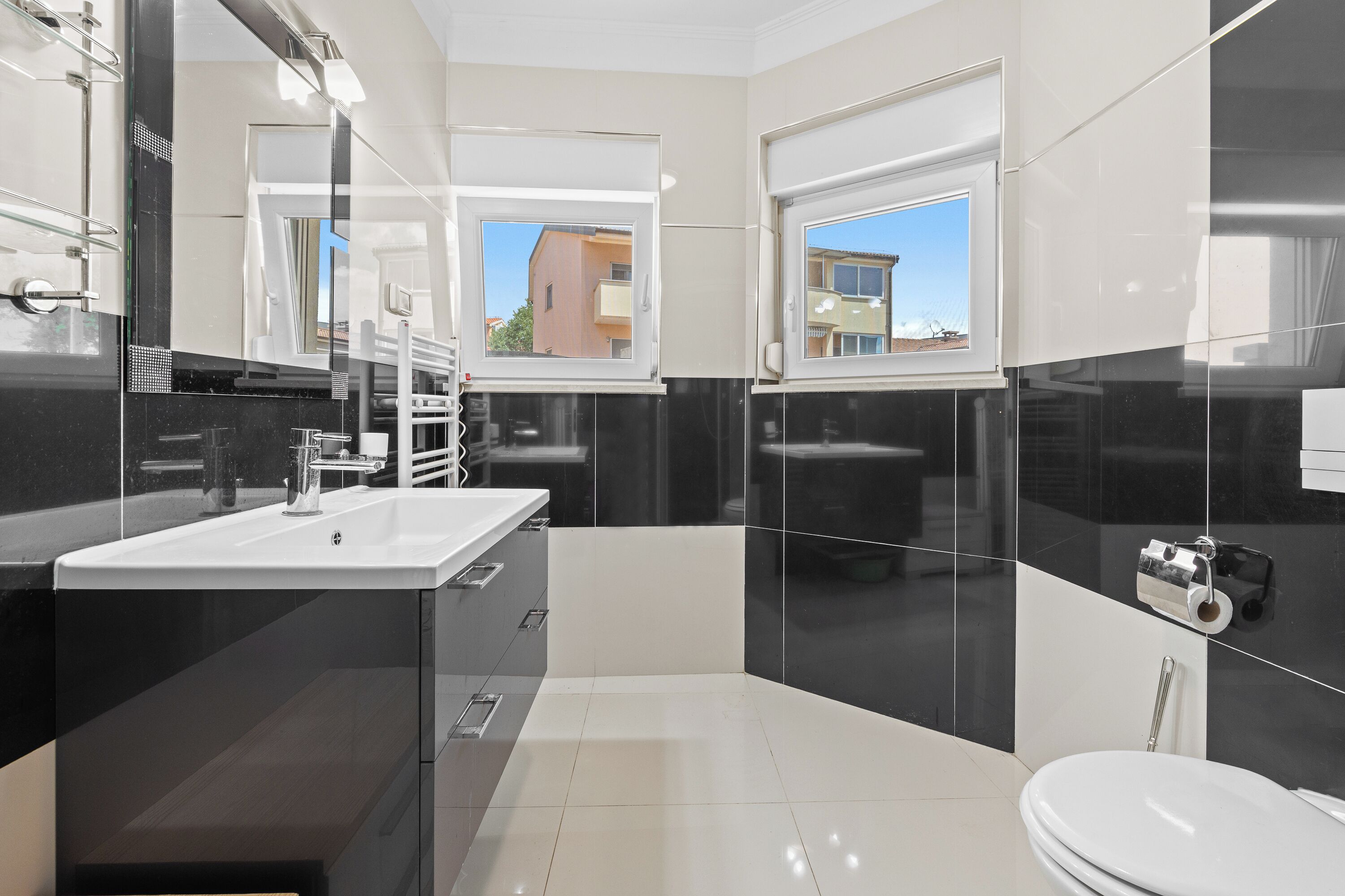 Un baño moderno con un lavamanos blanco, baldosas blanco y negro, un inodoro y una ventana que ofrece vistas a edificios.