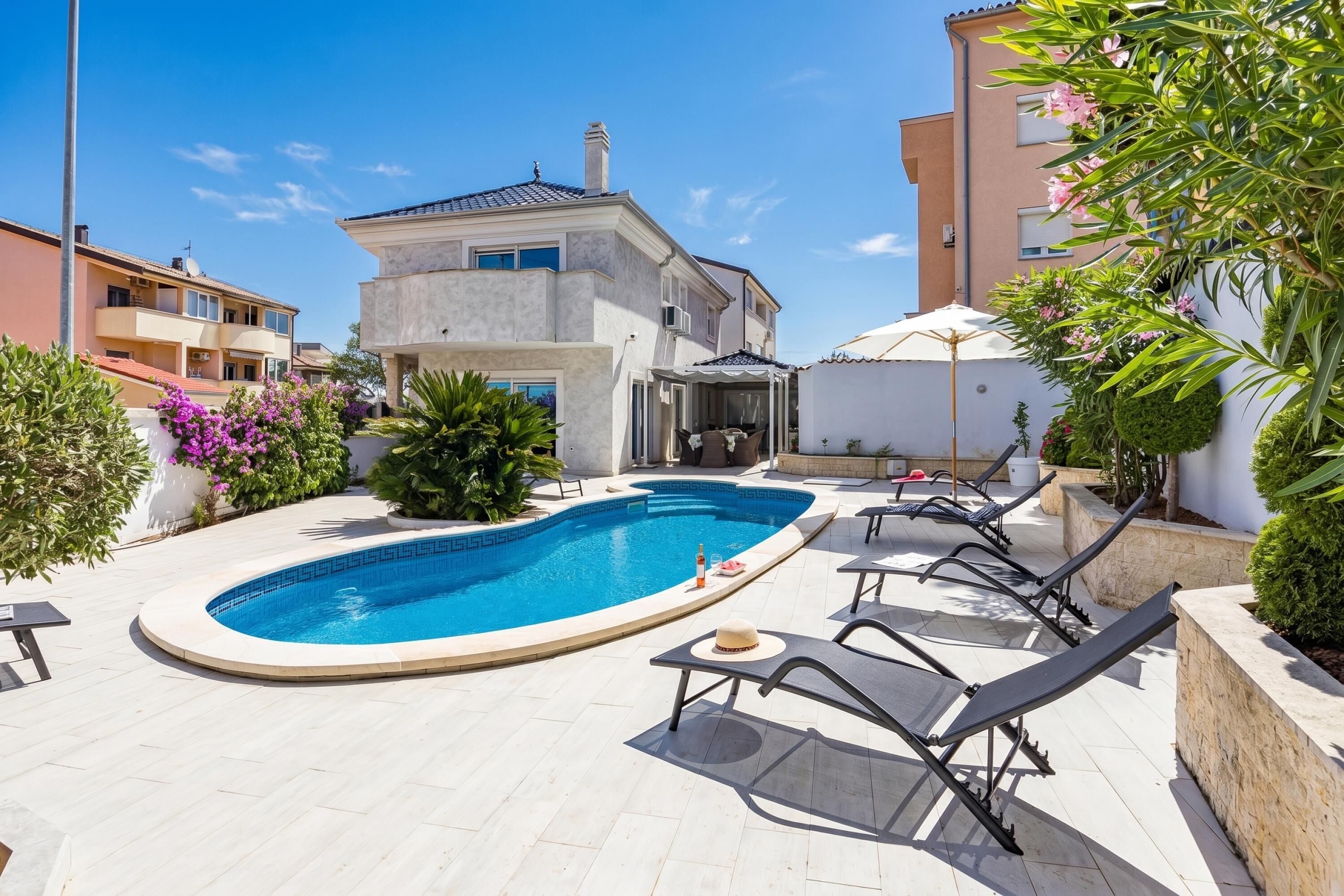 A swimming pool with lounge chairs and a sun umbrella, surrounded by buildings and greenery.