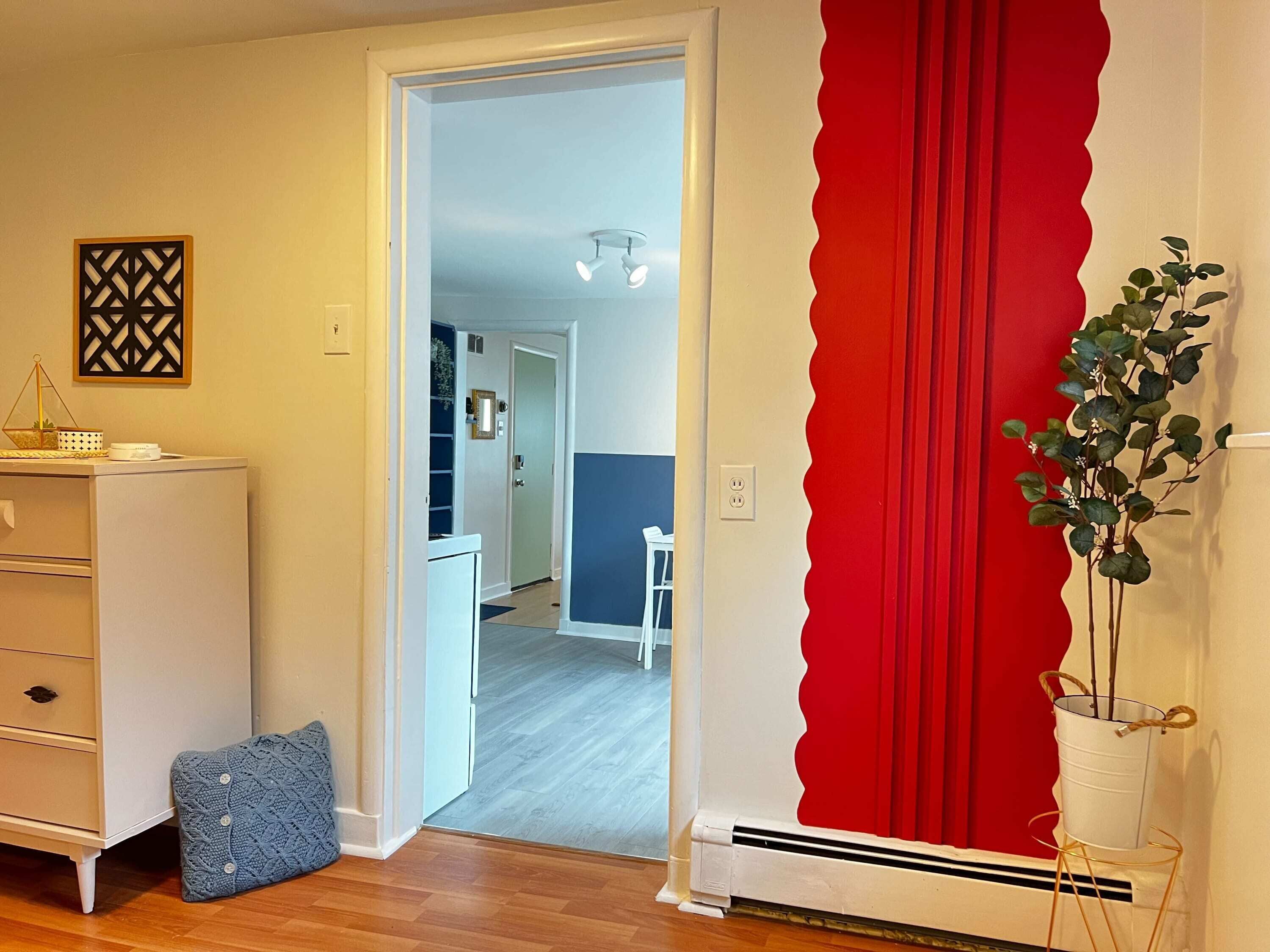 Una habitación con un aparador blanco, un otoman tejido azul, una pared de acento roja y una planta en una maceta blanca.