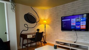 Ein modernes Zimmer mit Fernseher, der Roku anzeigt, einem Schreibtisch mit Stuhl, einem weißen Couchtisch und einer Lampe.