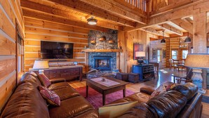 A log cabin interior with a stone fireplace, leather sofas, wooden floors, and a dining area. - Grand Vista Lodge (West Jefferson)