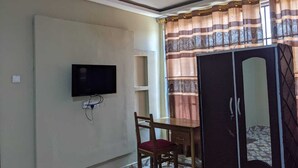 Ein Zimmer mit Fernseher an der Wand, einem Holztisch mit Stuhl, einem Kleiderschrank und einem Fenster mit Vorhängen.