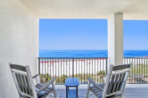 A balcony with two wooden chairs and a small table, overlooking a beach with umbrellas and a clear blue sky. - Windward Pointe 605 by Liquid Life (Orange Beach)
