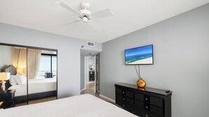 A hotel room with a bed, a dresser, a ceiling fan, a painting of a beach, and a view of the balcony. - Windward Pointe 1405 by Liquid Life (Orange Beach)