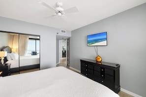 Habitación de hotel con cama, cómoda, ventilador de techo, pintura de una playa y vista al balcón.