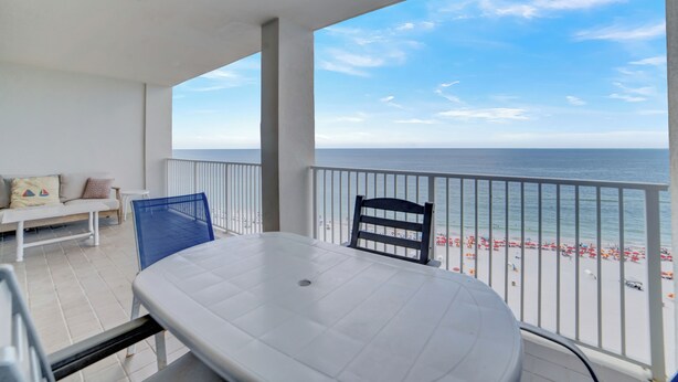 A balcony with a white table and chairs, a sofa, and a view of the beach and ocean. - Windward Pointe 1405 by Liquid Life (Orange Beach)