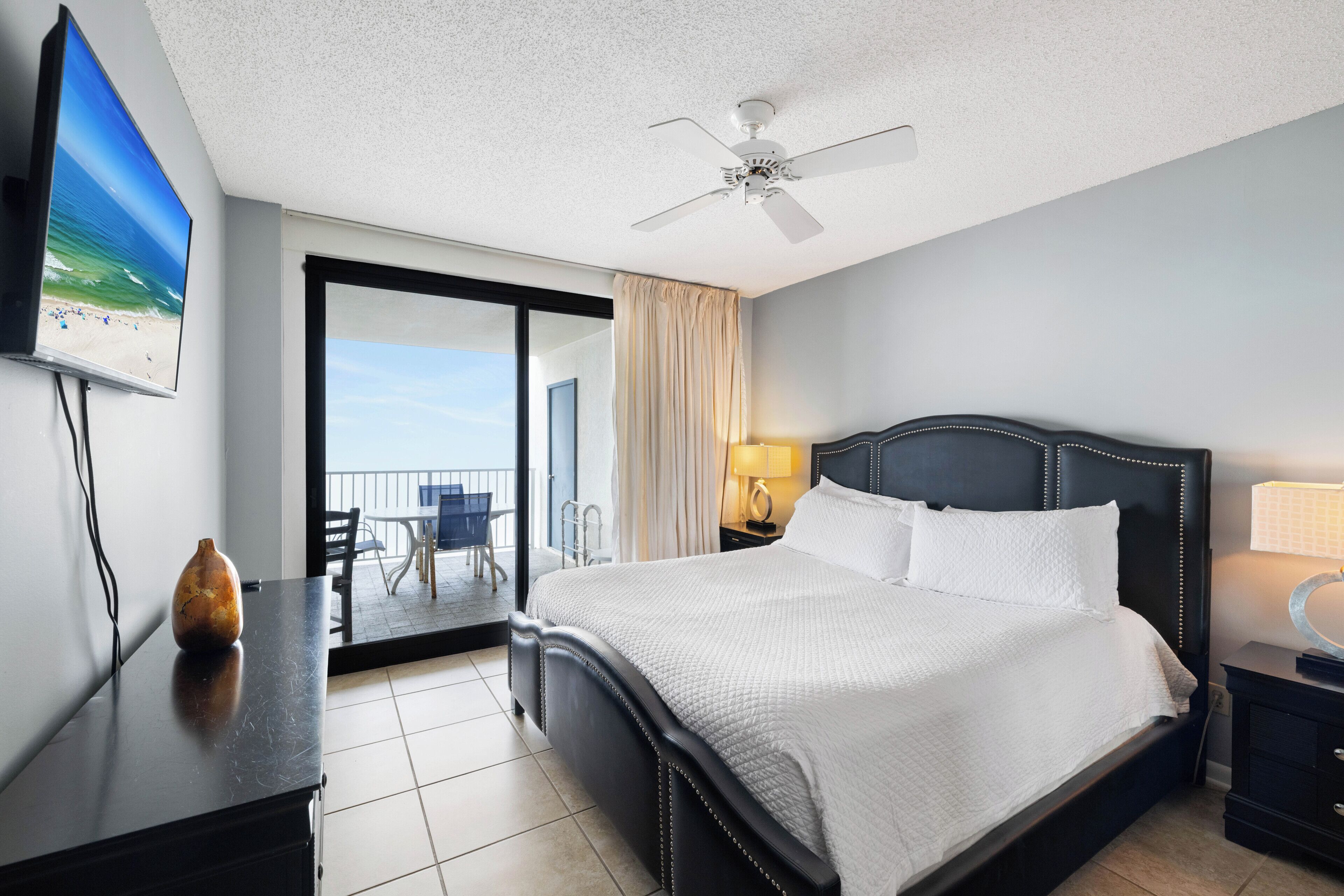 Une chambre d’hôtel avec un lit, un ventilateur de plafond, une télévision, un bureau avec un vase et une vue sur le balcon.