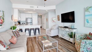 Una sala de estar moderna con un sofá, una mesa de centro y un televisor. Se observa una zona de cocina con armarios blancos y una mesa de comedor.