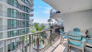 Un appartement moderne doté d’un balcon donnant sur une piscine et des palmiers, et comprenant un coin repas avec des chaises bleues et une table.