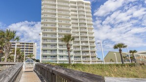 A tall, multi-story hotel with balconies, a wooden boardwalk, and palm trees. - Tradewinds 1207 by Liquid Life (Orange Beach)