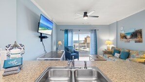 A kitchen with a double sink, a ceiling fan, a TV mounted on the wall, and a view of the outside through a window. - Tradewinds 1207 by Liquid Life (Orange Beach)