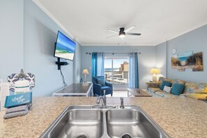 A kitchen with a double sink, a ceiling fan, a TV mounted on the wall, and a view of the outside through a window. - Tradewinds 1207 by Liquid Life (Orange Beach)