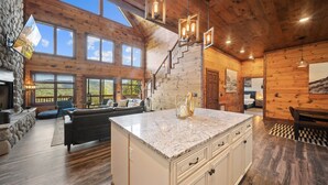 A spacious log cabin interior with a large kitchen island, a stone fireplace, and a cozy seating area with a sofa and chairs. - Cranberry View Lodge (Newland)