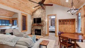 A cozy living room with a stone fireplace, a ceiling fan, and a dining area with wooden furniture. - Banner Bear (Banner Elk)