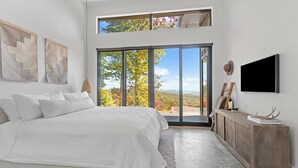 Ein modernes Schlafzimmer mit einem großen Bett, einer Schiebeglastür, die auf einen Balkon führt, und einem an der Wand befestigten Fernseher.