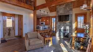 A spacious living area with a high ceiling, wooden beams, a stone fireplace, a dining table with chairs, and a kitchen with wooden cabinets and a microwave. - Mio Nido at Eagles Nest (Banner Elk)