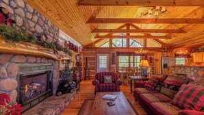A cozy log cabin interior with a stone fireplace, wooden ceiling beams, a red plaid sofa, and a dining area. - A Moon River View (Boone)