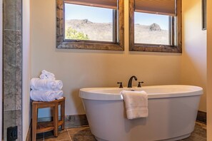 Une salle de bain avec une baignoire sur pieds, deux fenêtres à cadre en bois et une pile de serviettes blanches posée sur un tabouret en bois.