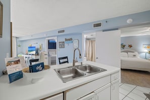 Une cuisine compacte dotée d’un évier à deux bacs, d’un plan de travail blanc et donnant sur une chambre à coucher avec un lit et une table de chevet.