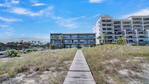 Een strandhotel met een promenade die naar het gebouw leidt, meerdere balkons en palmbomen.