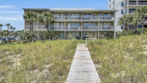 Een strandhotel met een houten promenade die naar de ingang leidt, meerdere balkons en een parkeerplaats op de achtergrond.