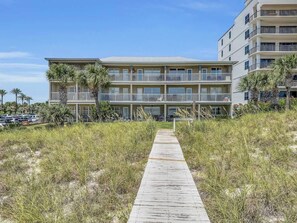 Ein Hotel am Strand mit einer Holzpromenade, die zum Eingang führt, mehreren Balkonen und einem Parkplatz im Hintergrund.