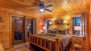 A wooden cabin bedroom with a bed, two bedside tables, a ceiling fan, and a window with curtains. - Bearadise at Blue Ridge (West Jefferson)