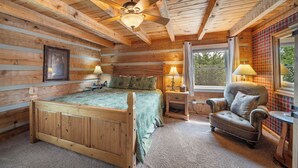 A wooden cabin bedroom with a bed, two bedside lamps, a chair, and a window with curtains. - Napping Bear Cabin (Boone)