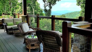 A wooden deck with wicker furniture and a glass table, overlooking a lush green landscape with mountains in the distance. - Heritage Lodge at Eagles Nest (Banner Elk)
