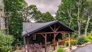 A rustic cabin with a stone foundation, wooden porch, and a metal roof, surrounded by lush greenery and trees. - Sky Laurel Cottage (Blowing Rock)