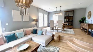 A modern living room with a large sectional sofa, a wooden coffee table, and a dining area with a chandelier. - The Cliffs - Sabaido Mare (Manón)