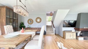 A dining area with a wooden table, cushioned chairs, and a chandelier. There is a staircase leading to an upper level with a TV and fireplace. - The Cliffs - Sabaido Mare (Manón)