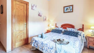 Un dormitorio con puerta de madera, una cama con colcha azul estampada, dos mesitas de noche con lámparas y tres cuadros enmarcados en la pared.