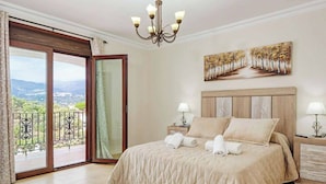 A bedroom with a large bed, two bedside tables, a chandelier, a painting, and a balcony with a view. - Villa Lara Pinta (Nerja)