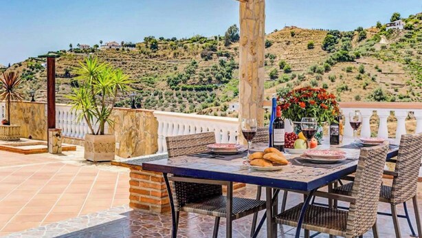 Un espace de restauration en plein air avec une table dressée pour deux personnes, entouré de chaises en osier, offrant une vue sur un paysage vallonné.