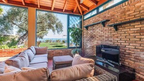 A cozy living area with a brick fireplace, comfortable seating, and large windows offering a view of the outdoors. - Villa Barbati Beachfront (Corfu)