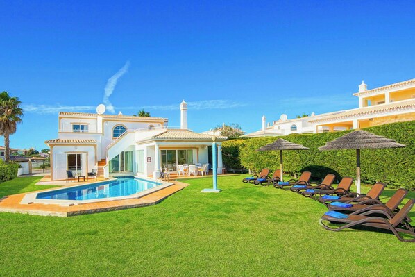 A poolside area with lounge chairs, a swimming pool, and a building with a white facade and a tall tower. - Villa Beatrice (Albufeira)
