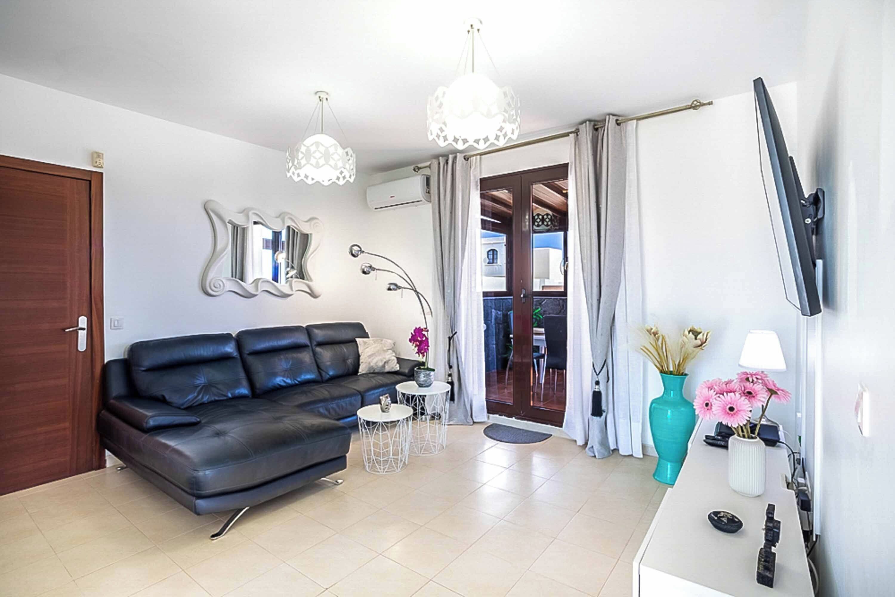 A modern living room with a black leather sofa, two small side tables, a large mirror, a flat-screen TV, and a teal vase with flowers.