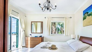 A bedroom with a bed, a dresser, a mirror, a chandelier, and a window with curtains. - Villa Julieta (Albufeira)