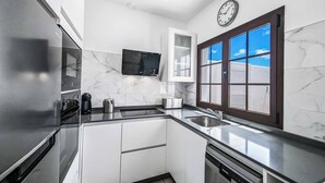 A modern kitchen with a stainless steel refrigerator, a built-in oven, a microwave, a coffee maker, and a dishwasher. There is a window with a view of the sky and a clock on the wall. - Villa Emilia (Yaiza)