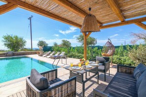 A poolside area with a wooden pergola, lounge chairs, and a table set for two with a basket and fruit.