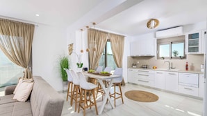 A modern kitchen with white cabinets, a dining area with a white table and chairs, and a living area with a sofa and curtains. - Villa Elios Residence (Rhodes)