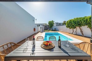 A poolside dining area with a table set for two, a bottle of wine, and a fruit bowl.