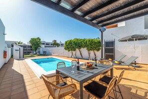 A rooftop pool area with a table set for dining, surrounded by lounge chairs and a pergola.