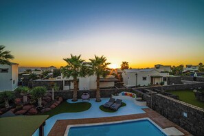 A rooftop pool area with lounge chairs and palm trees, surrounded by modern buildings during sunset.