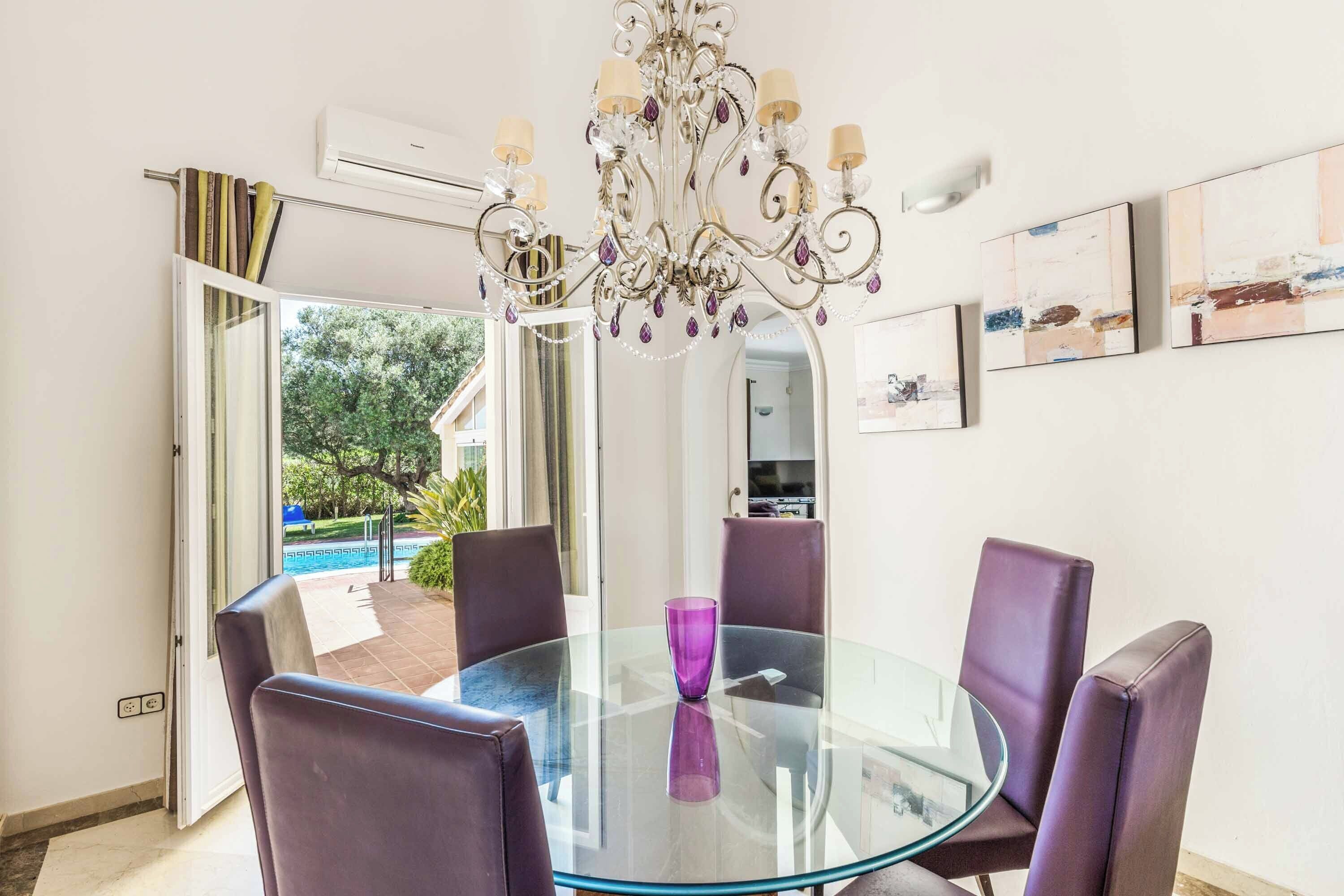 A dining area with a glass table, purple chairs, a chandelier, and a view of a pool.