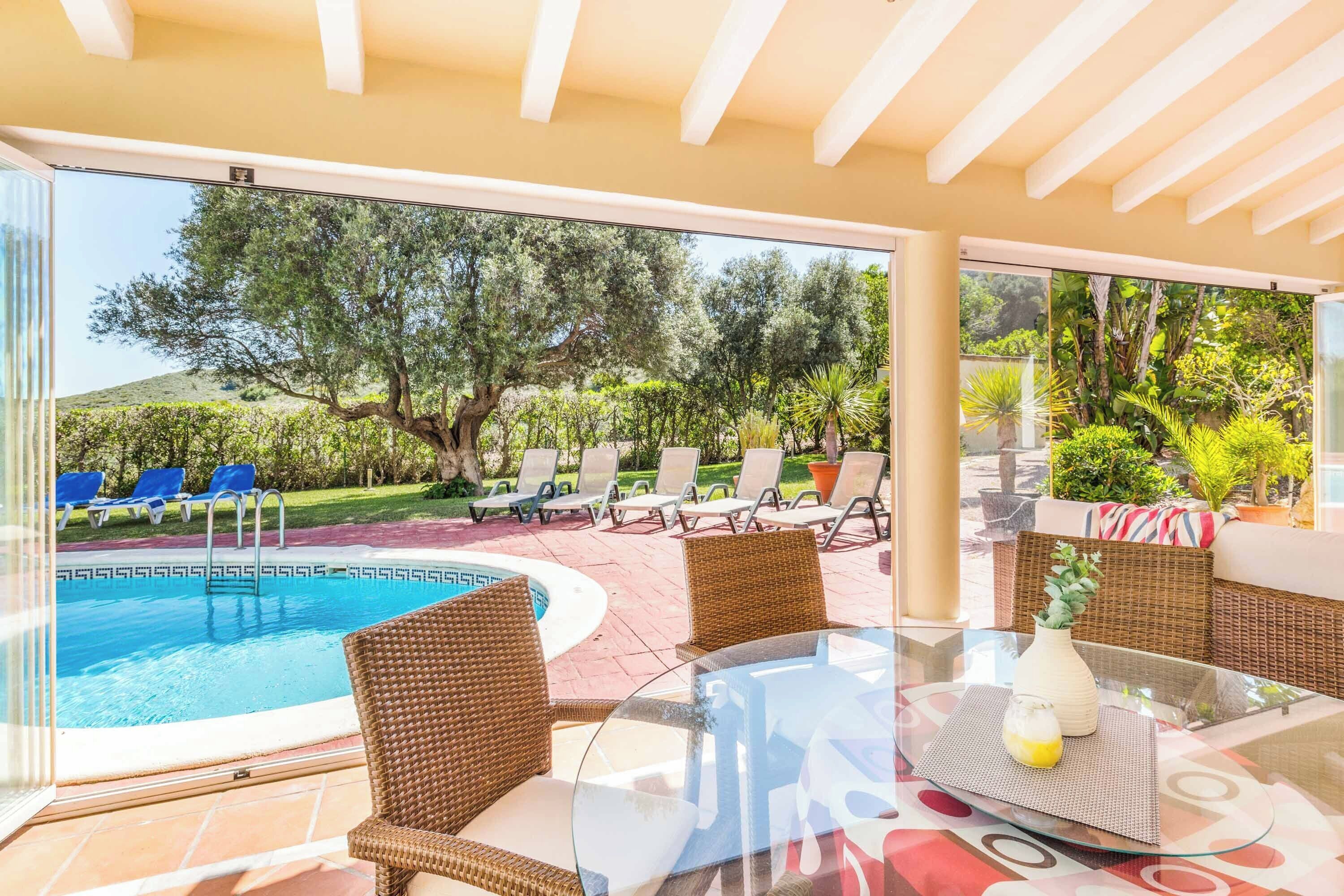 A poolside area with wicker chairs and a glass table, overlooking a swimming pool and a landscaped garden.