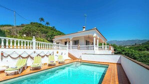 A pool area with lounge chairs, a white building with a red roof, and a view of hills and mountains. - Villa Carmela Nerja (Nerja)